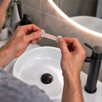 Hands holding a NasalFlow nasal strip by the white backing paper at each end, the sticky centre exposed and untouched