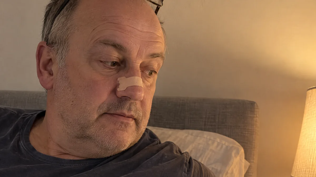 Man sitting up in bed beside a warm lamp, wearing a nasal strip across his nose